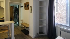 A compact living space with a wooden floor, a small table with a cup and remote, a bench, a white cabinet, and a window with a view of a stone wall.