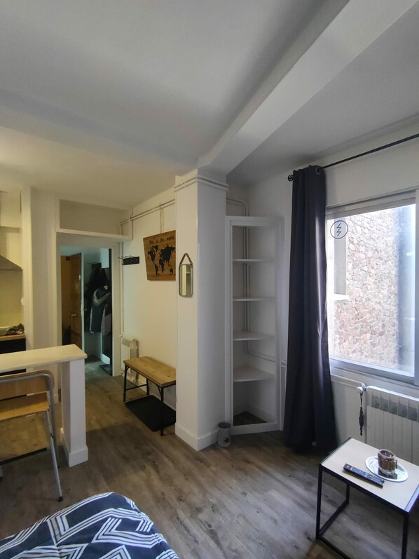 A compact living space with a wooden floor, a small table with a cup and remote, a bench, a white cabinet, and a window with a view of a stone wall.