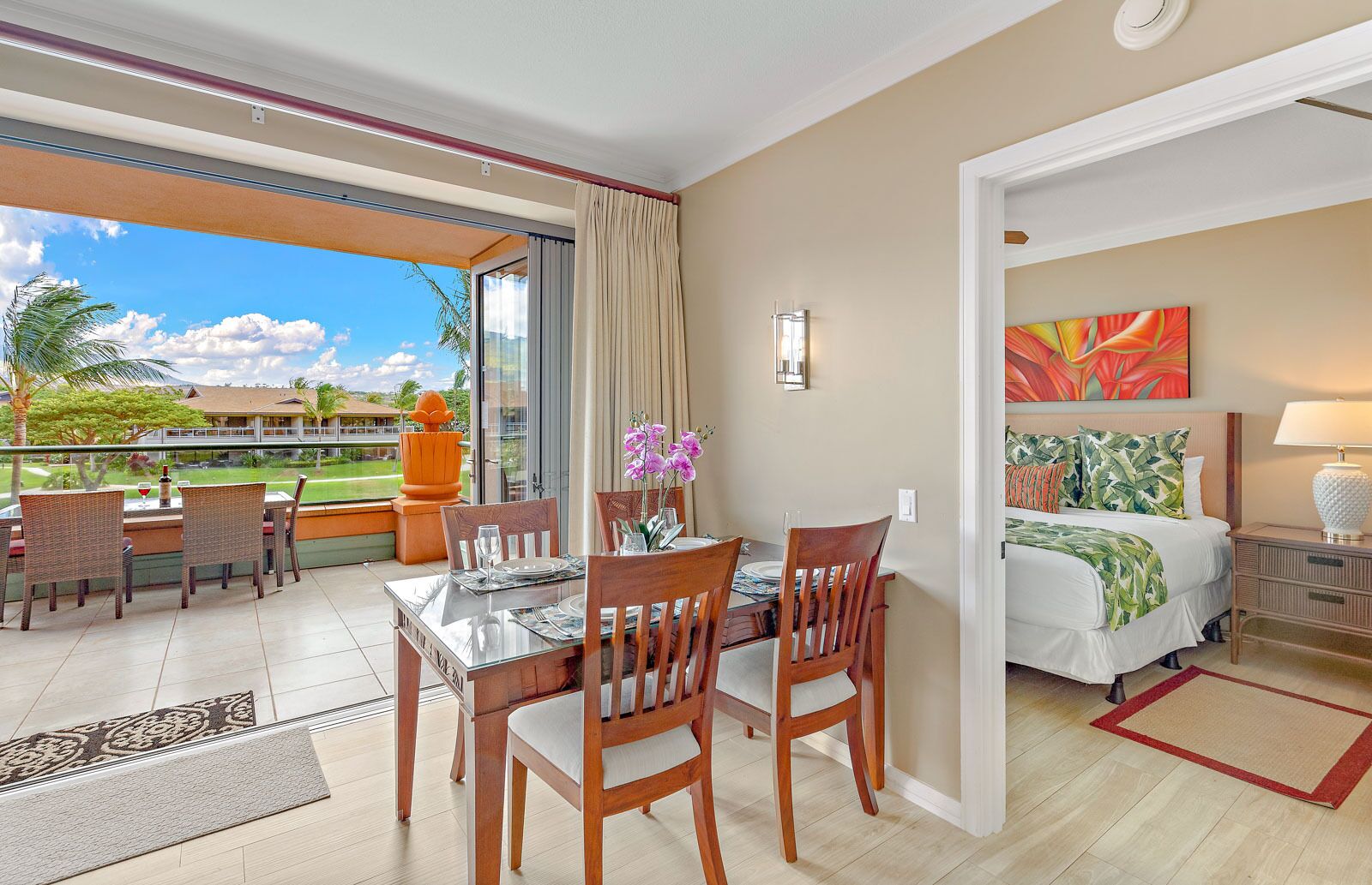 Habitación de hotel con balcón, zona de comedor con mesa y sillas, cama con colcha de motivos tropicales y un cuadro en la pared.