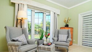 A room with two armchairs, a small ottoman, and a lamp. There is a window with curtains and a view of greenery outside.