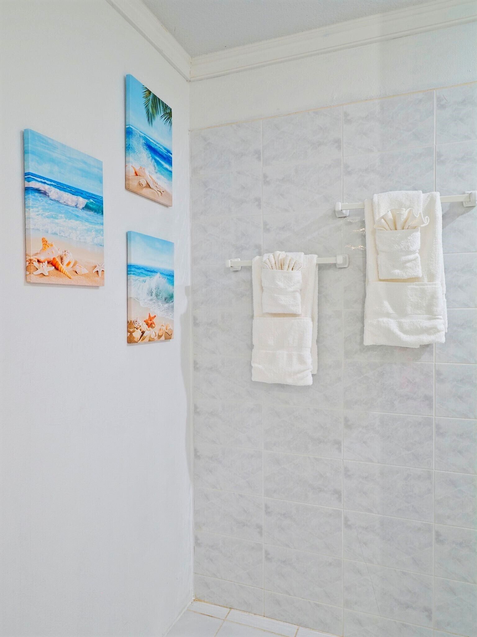 Une salle de bain aux carreaux blancs, avec deux serviettes blanches suspendues à un porte-serviettes et trois tableaux sur le thème de la plage accrochés au mur.