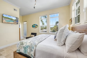 A bedroom with a bed, a painting of a pool, and a window with a view of palm trees.