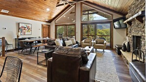 Amplia sala de estar con techo alto, vigas de madera, chimenea y ventanas grandes que ofrecen vistas al exterior.