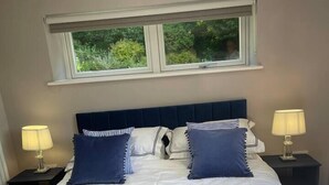 A neatly made bed with a dark headboard, two bedside lamps, and a window with a view of greenery. - Brighouse Studio - Pet Friendly! - Free Parking (Brighouse)
