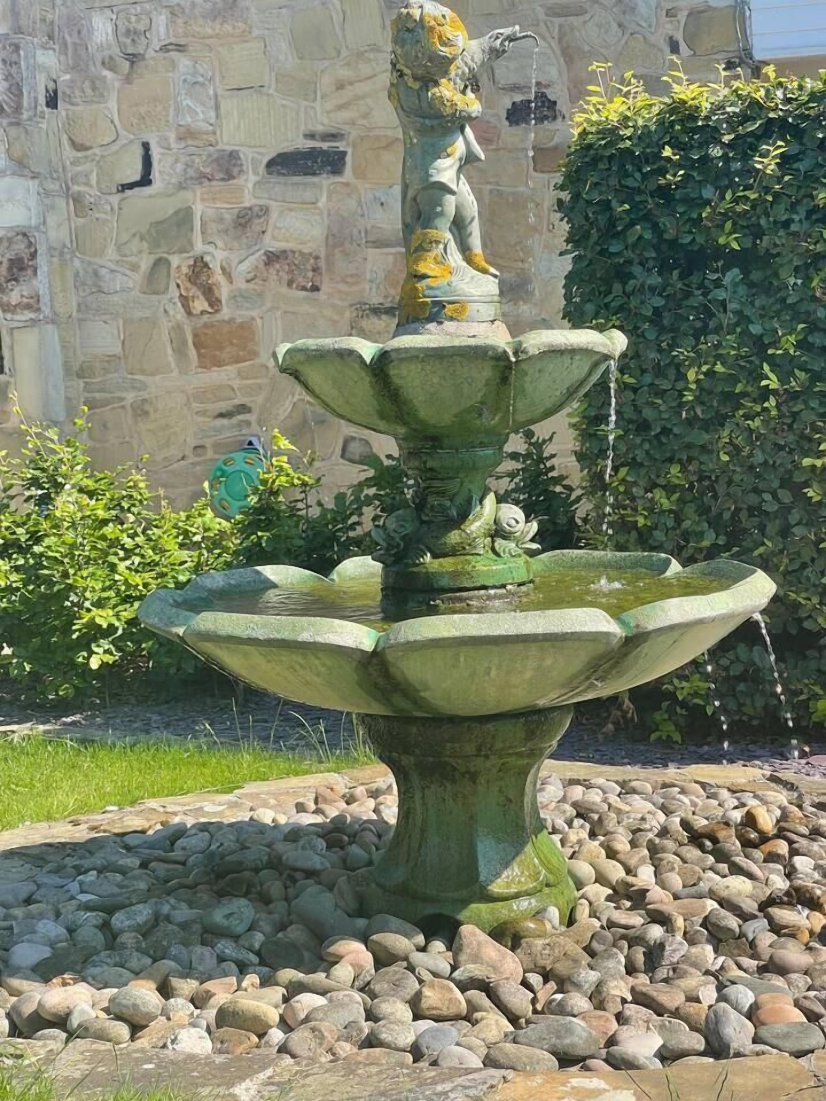 A stone fountain with a statue of a figure pouring water, surrounded by a bed of rocks and greenery.