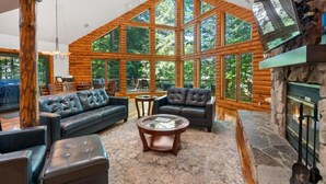 A log cabin interior with a stone fireplace, leather sofas, and large windows offering a view of the forest.