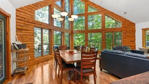 A log cabin interior with a dining area, a sofa, and large windows offering a view of the outdoors.