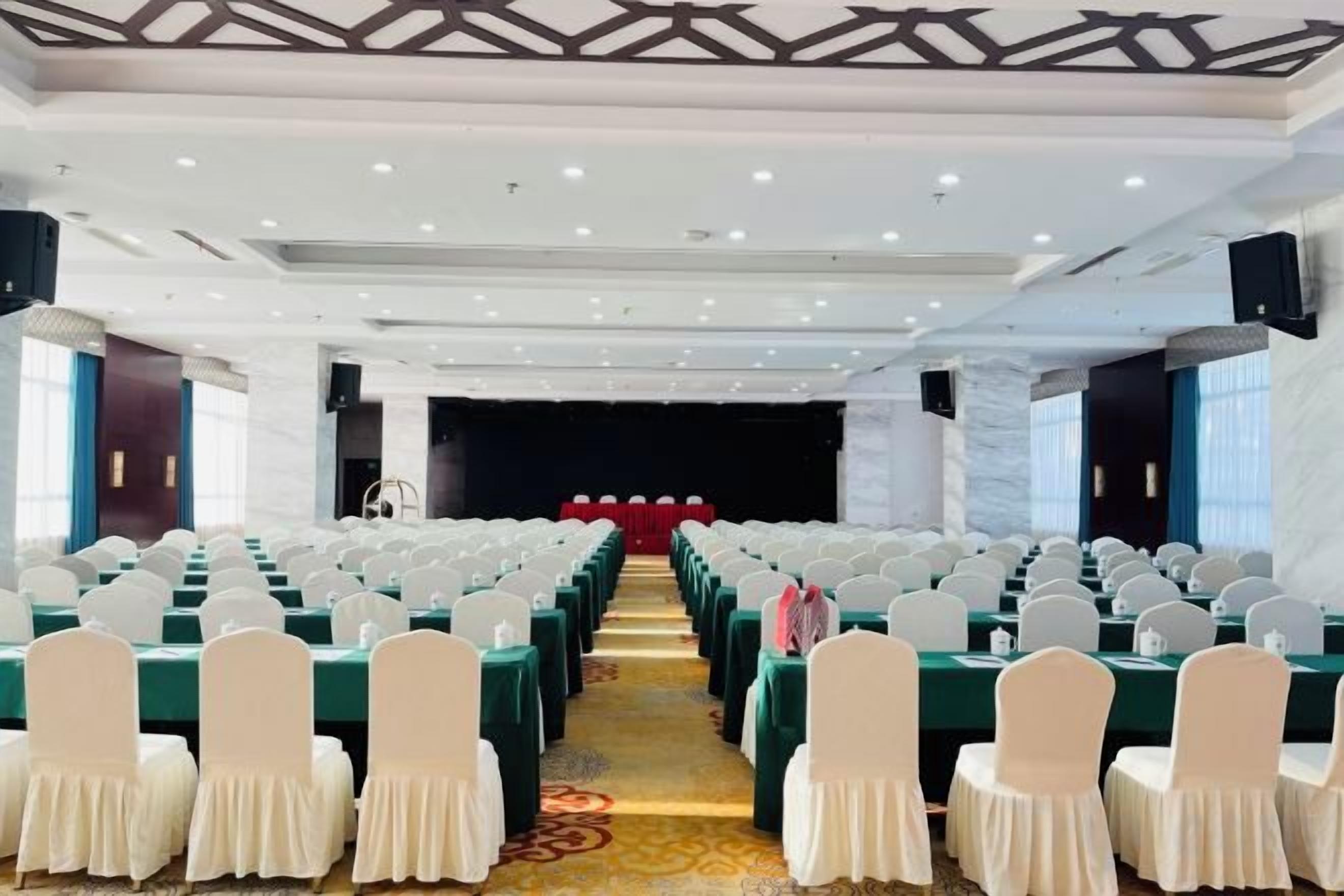 Un salón de conferencias con filas de mesas y sillas, un escenario y suelo alfombrado.