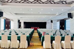 Un salón de conferencias con filas de mesas y sillas, un escenario y suelo alfombrado.