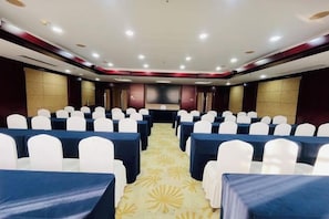 A conference room with rows of tables and chairs, a stage, and a carpeted floor.