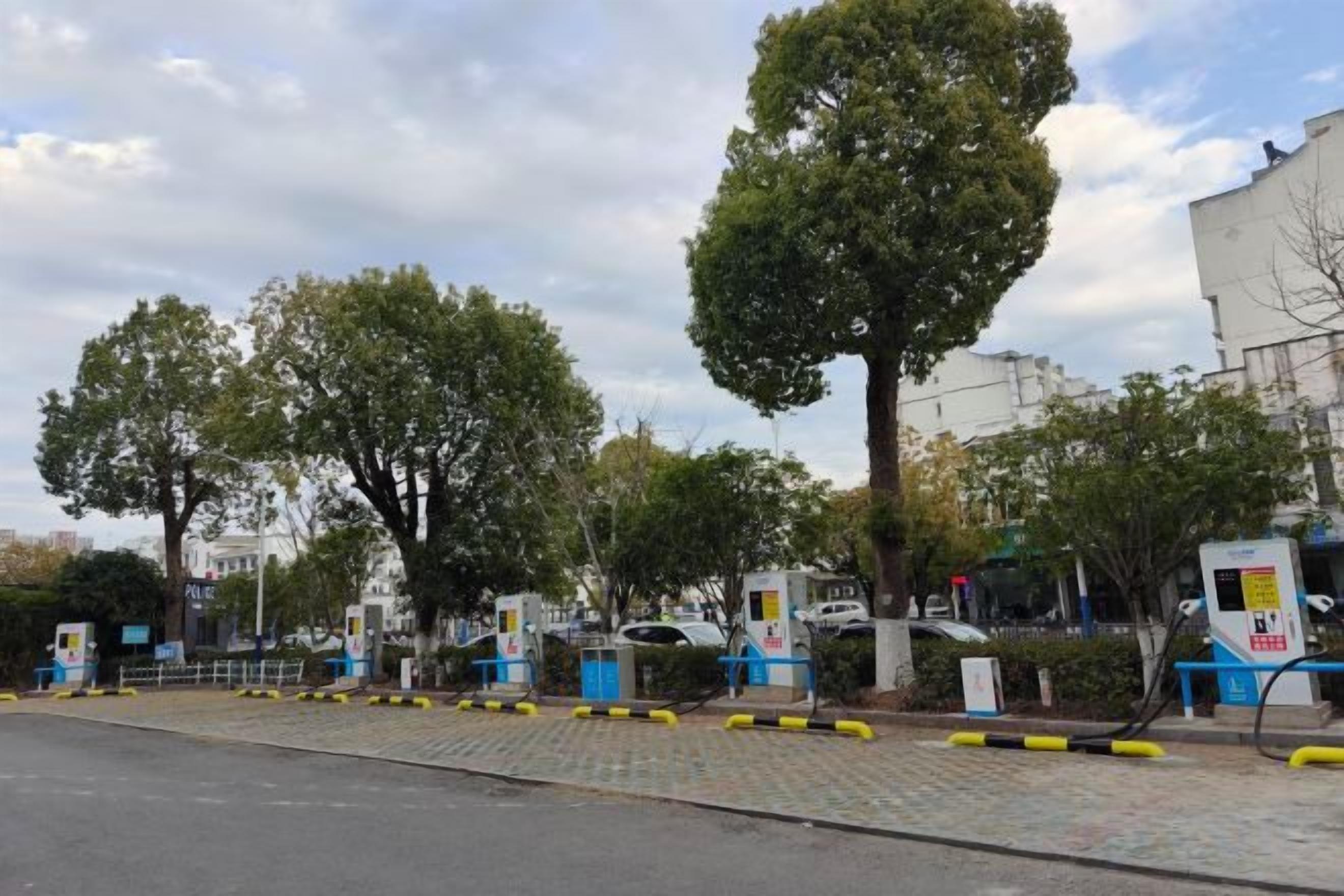 A parking area with electric vehicle charging stations, trees, and a building in the background.
