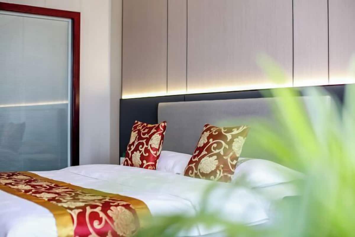 A hotel room with a bed, a window with blinds, and a headboard with built-in lighting.