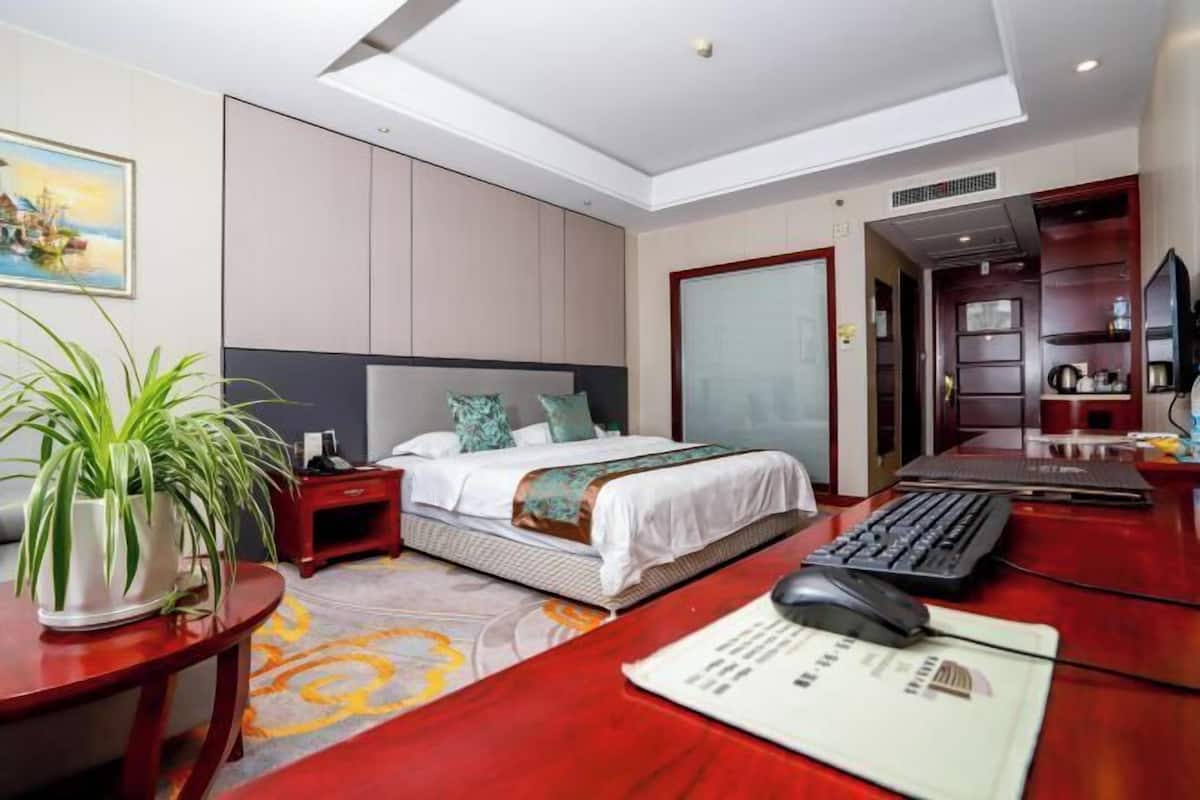 A hotel room with a bed, desk, computer, and a potted plant.