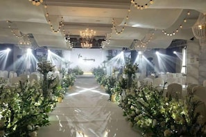An indoor venue with a central aisle flanked by floral arrangements, a chandelier, and bright overhead lighting.