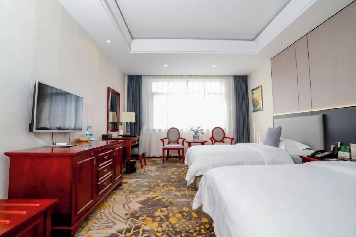 A hotel room with two beds, a TV, a desk, a red dresser, and a window with curtains.