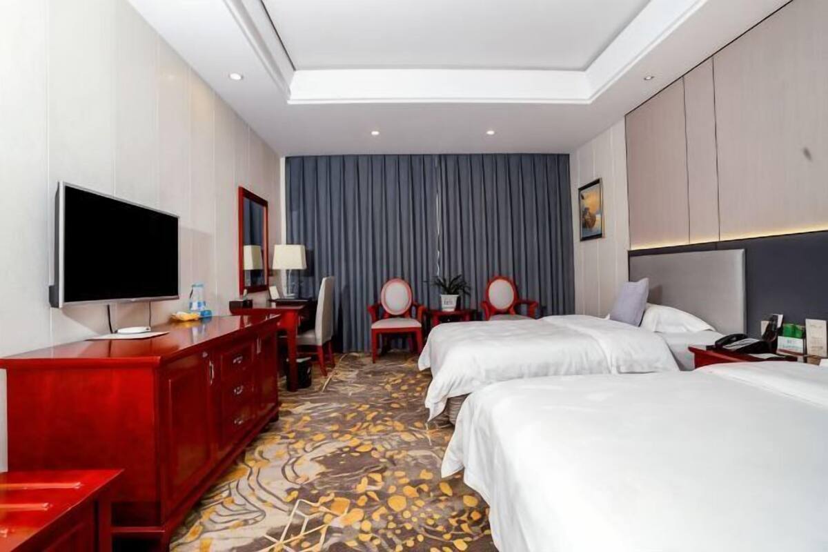 A hotel room with two beds, a TV, a desk, a chair, and a large window with curtains.