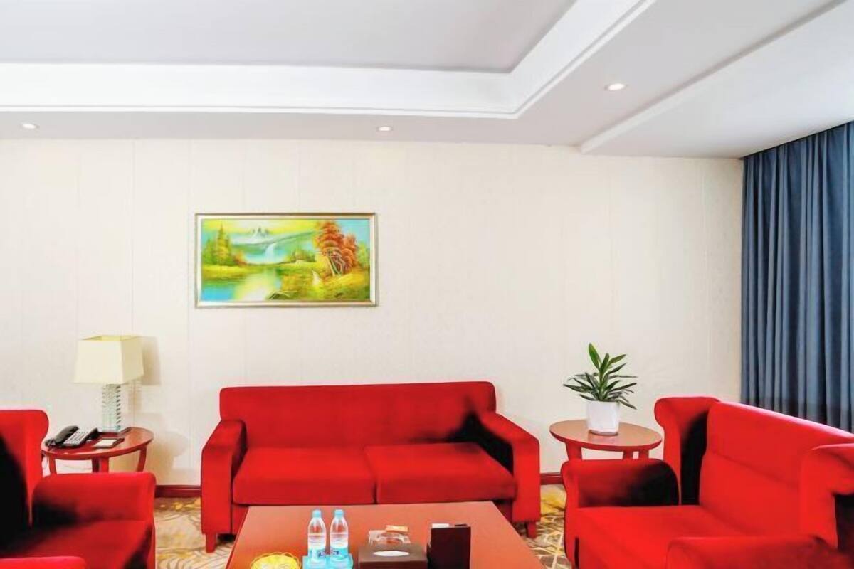 A hotel room with a red sofa set, a small table with a lamp, a painting on the wall, and a potted plant.