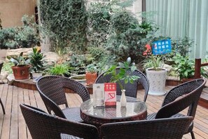 Een buitenplaats met rieten stoelen en een ronde tafel, omgeven door potplanten en een bord met tekst.