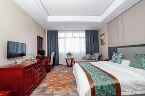 Quarto de hotel com cama, televisão, escrivaninha, cadeira, planta e janela com cortinas.