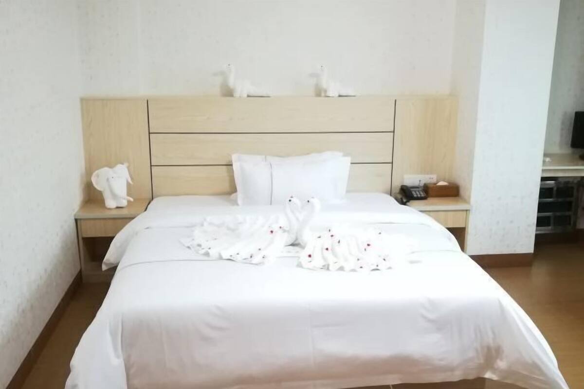 Een net opgemaakt bed met witte lakens en decoratieve zwanen, een houten hoofdbord en een klein nachtkastje met een witte beeldfiguur.