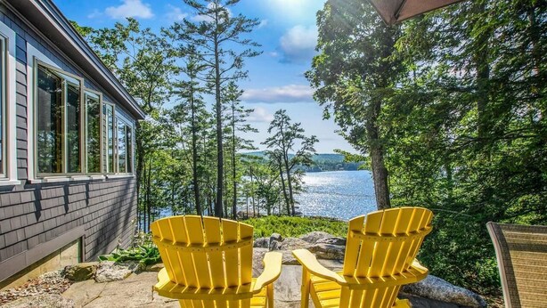 A patio with Adirondack chairs overlooking a lake, a wooden deck, and a house with large windows. - Webster Lake Retreat for Nature and Water Lovers (Franklin)