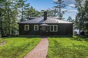 Une petite maison aux couleurs sombres, avec un chemin de briques menant à l’entrée, entourée d’une végétation luxuriante et d’arbres.