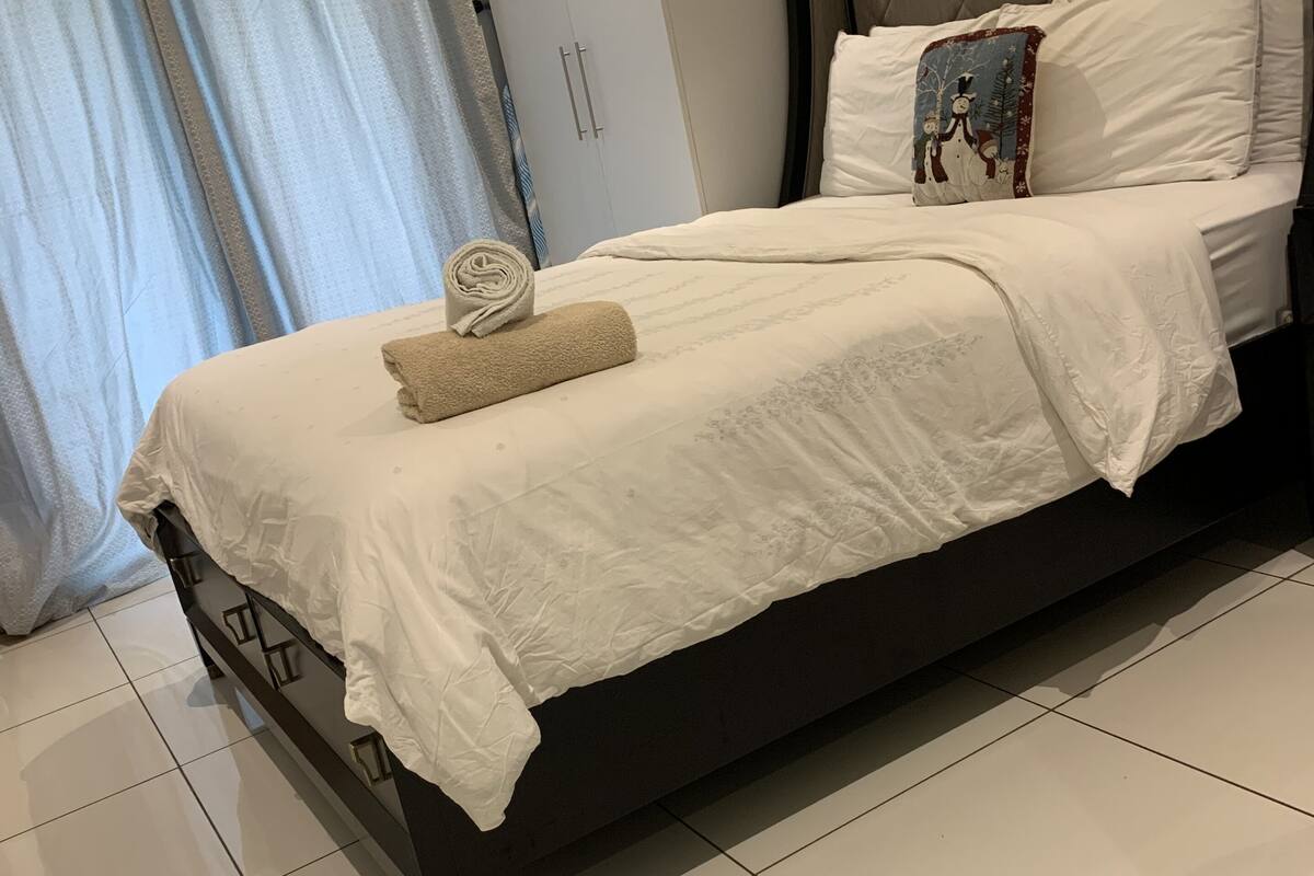 A neatly made bed with a rolled towel, a pillow with a winter-themed design, and a wardrobe in the background.