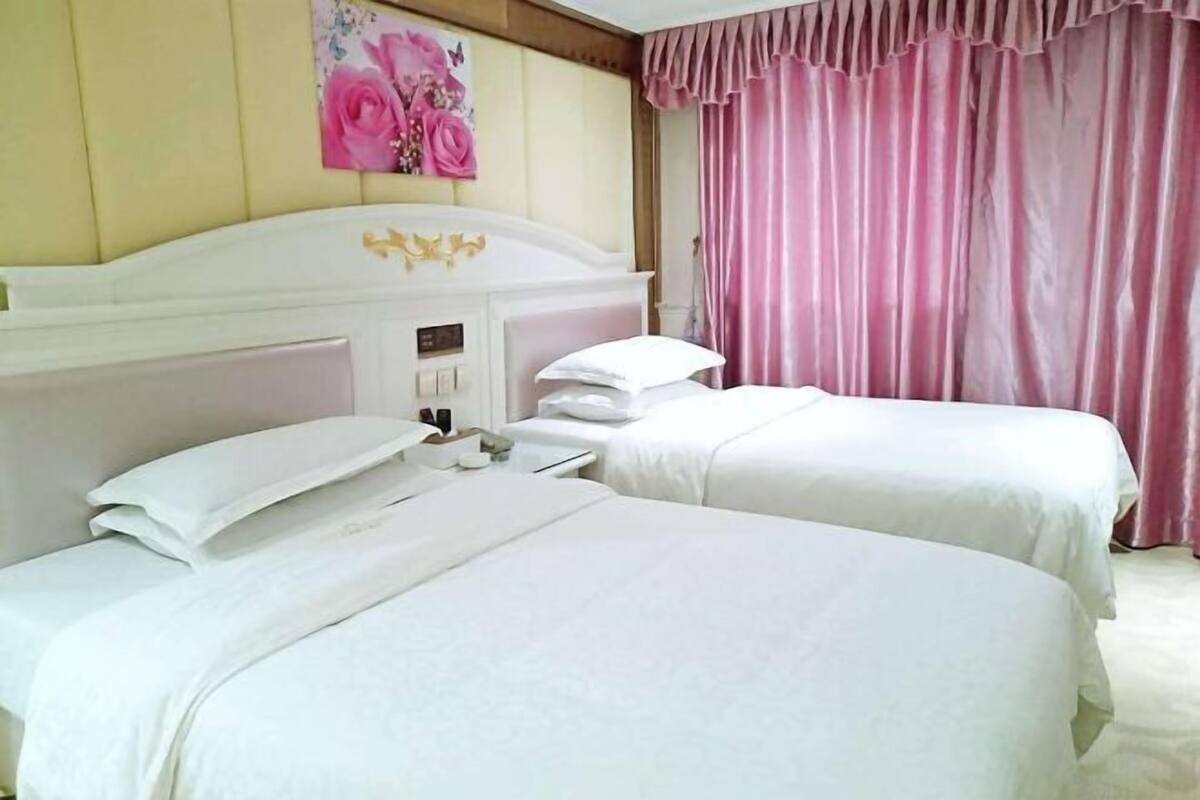 Habitación de hotel con dos camas, cabecera con reloj y cortina rosa.