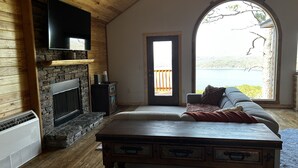 Una habitación acogedora con chimenea de piedra, televisor montado en la pared, un sofá cómodo y un amplio ventanal con vistas a un lago.