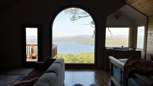 Una habitación con un amplio ventanal que ofrece vistas a un lago y colinas. En el interior, hay un sofá, un aparador de madera y una alfombra.