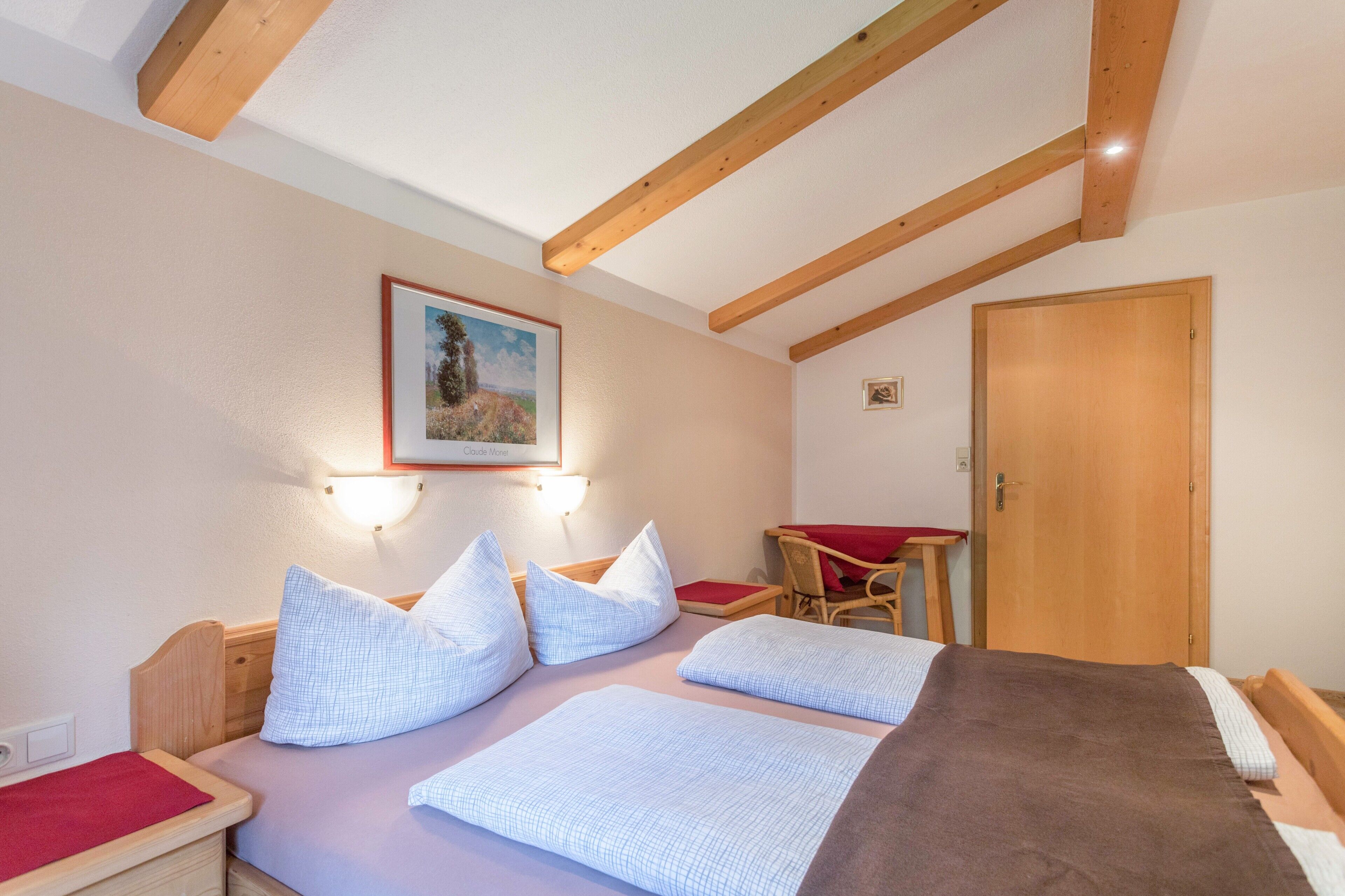 A bedroom with a bed, pillows, a wooden headboard, a wall-mounted light, a door, and a small table with a red tablecloth.