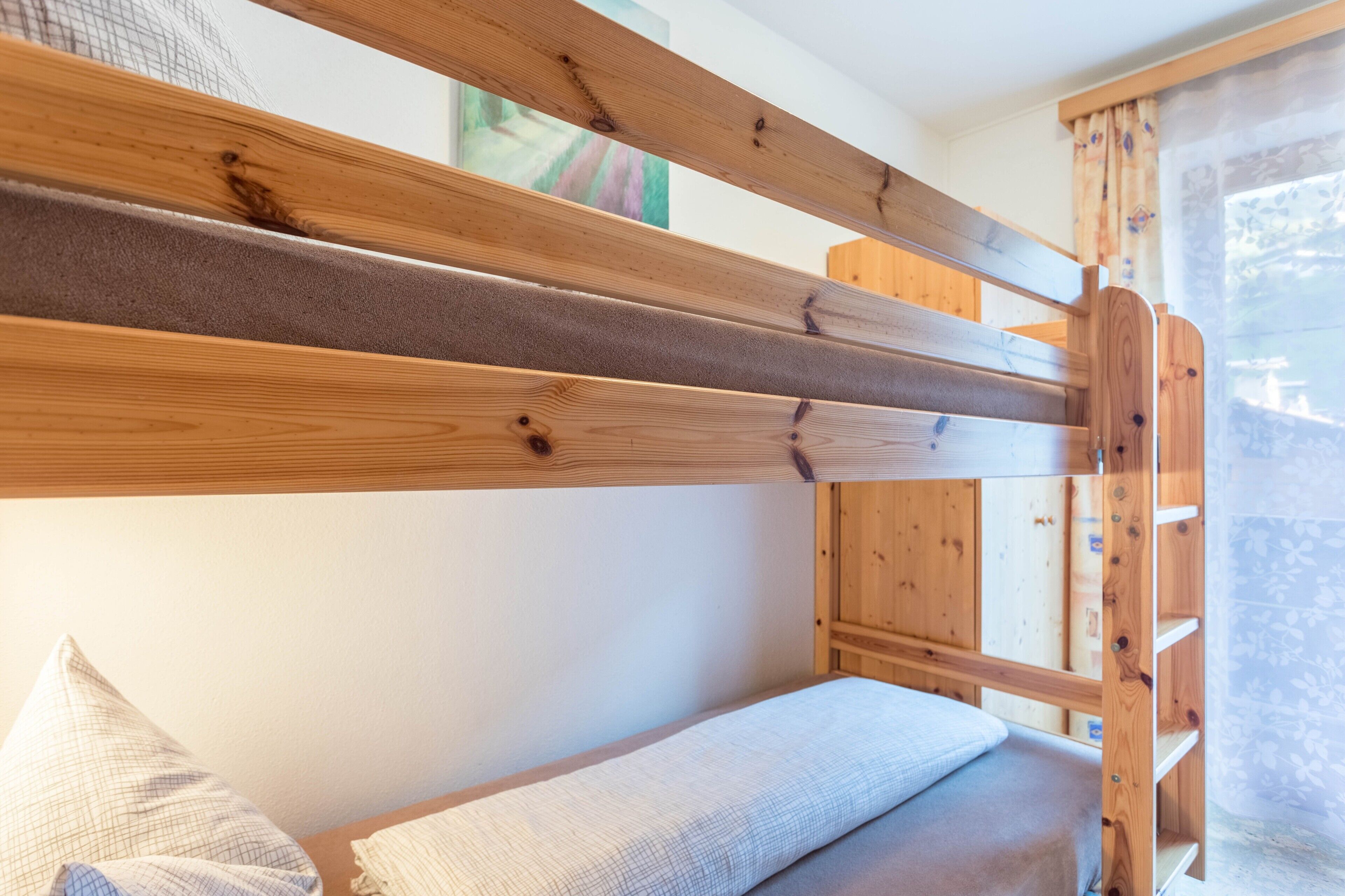 Una cama litera con listones de madera, un almohadón y una ventana con cortinas.