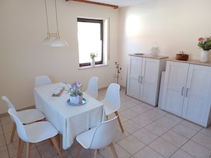Un comedor con una mesa dispuesta para cuatro personas, sillas blancas, una ventana con vistas y un mueble con objetos decorativos.