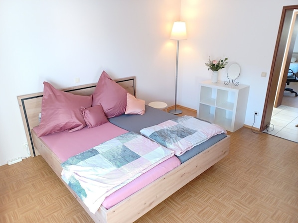 Una cama bien hecha con ropa de cama rosa y gris, una lámpara de noche, una mesita de noche pequeña blanca y un espejo en la pared.