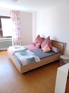 Une chambre à coucher avec un lit en bois, un radiateur, une fenêtre avec des rideaux et une petite table d’appoint ronde.