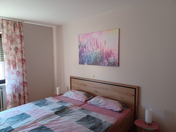 Une chambre avec un lit en bois, un rideau à motifs floraux, une couverture rose et grise, un tableau représentant des fleurs et une table de chevet avec une bougie.
