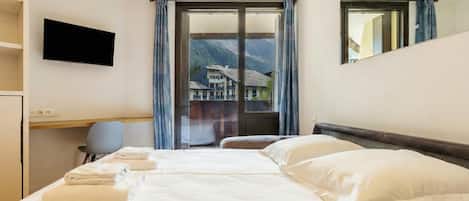 A hotel room with a large bed, a flat-screen TV mounted on the wall, a desk with a chair, and a view of mountains through the window.