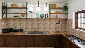 Una cocina con salpicadero de ladrillo, armarios de madera, un lavamanos de acero inoxidable y una estufa a gas. Hay luces colgantes y repisas con objetos decorativos.