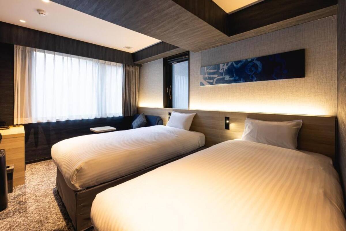 Una habitación de hotel con dos camas, un ventanal con cortinas, una obra de arte colgada en la pared y una mesita de noche.