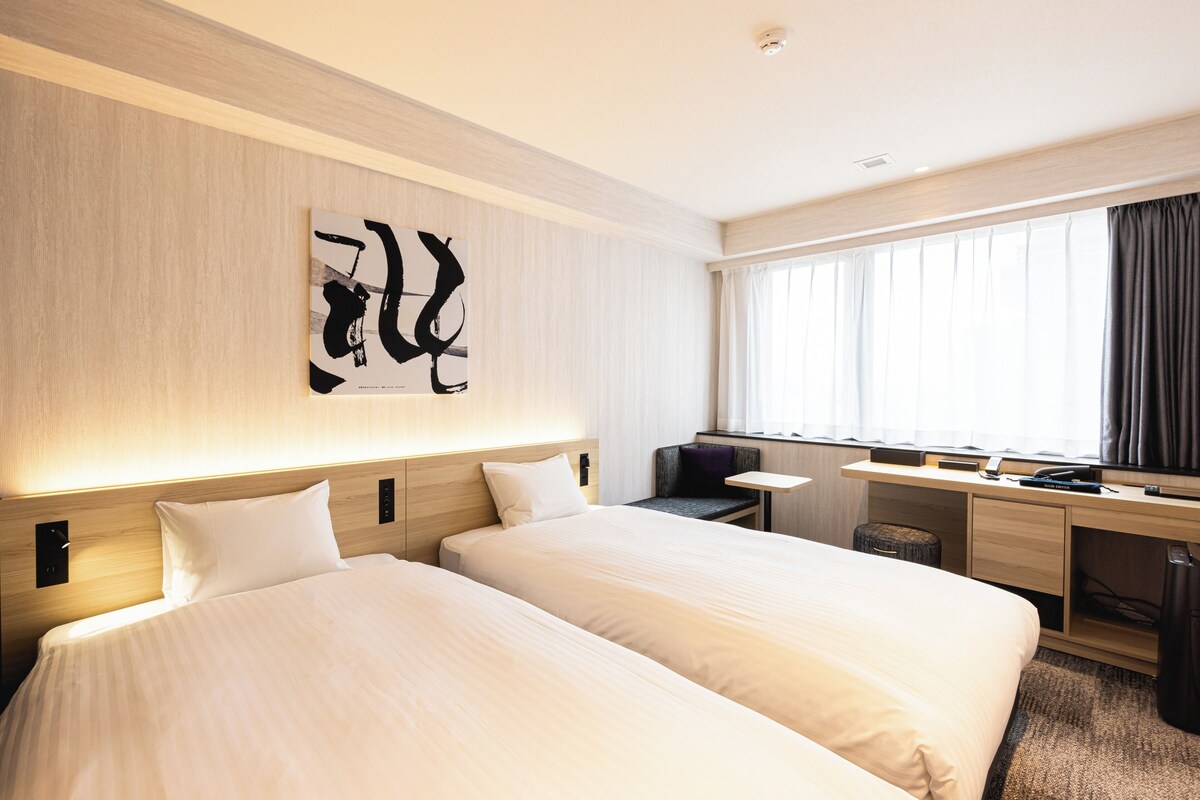 Habitación de hotel con dos camas, un escritorio, una silla, un ventanal con cortinas y una obra de arte en la pared.