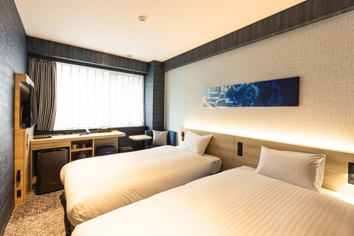 Una habitación de hotel con dos camas, un escritorio, una silla, un televisor y una ventana con cortinas.