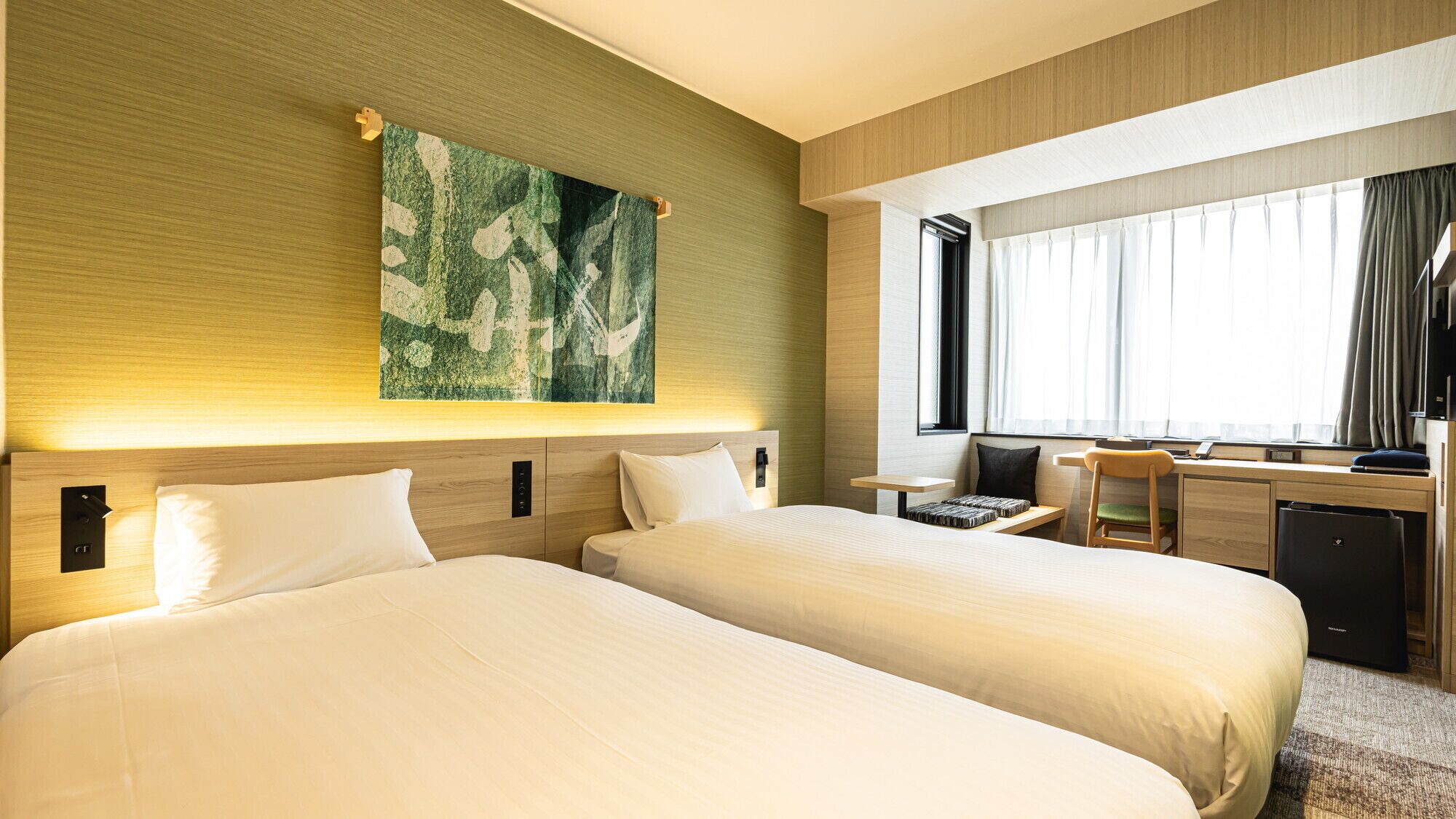 Quarto de hotel com duas camas, uma escrivaninha, uma cadeira e uma janela grande com cortinas.