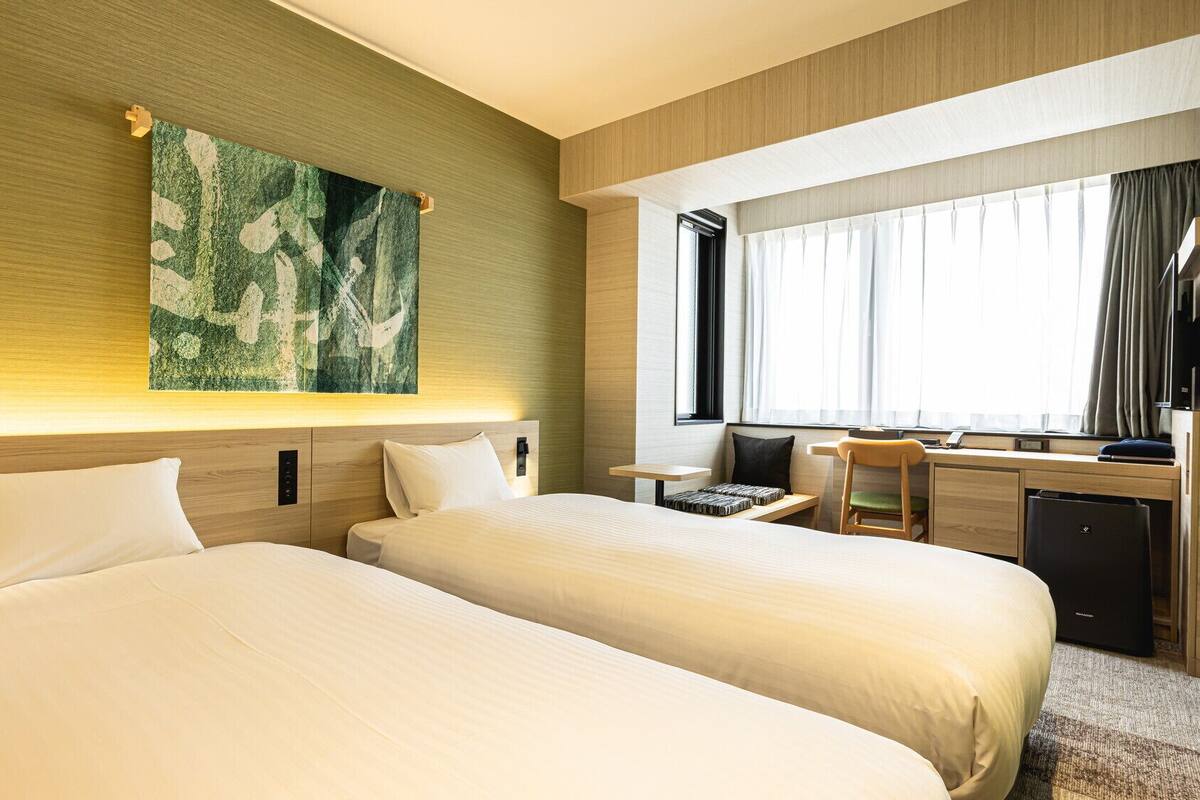 Habitación de hotel con dos camas, un escritorio, una silla y un ventanal con cortinas.