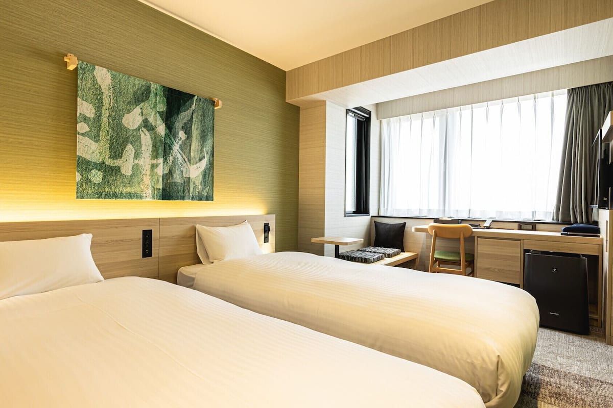 Hotellihuone, jossa on kaksi sänkyä, työpöytä, tuoli ja suuri ikkuna, jossa on verhot.