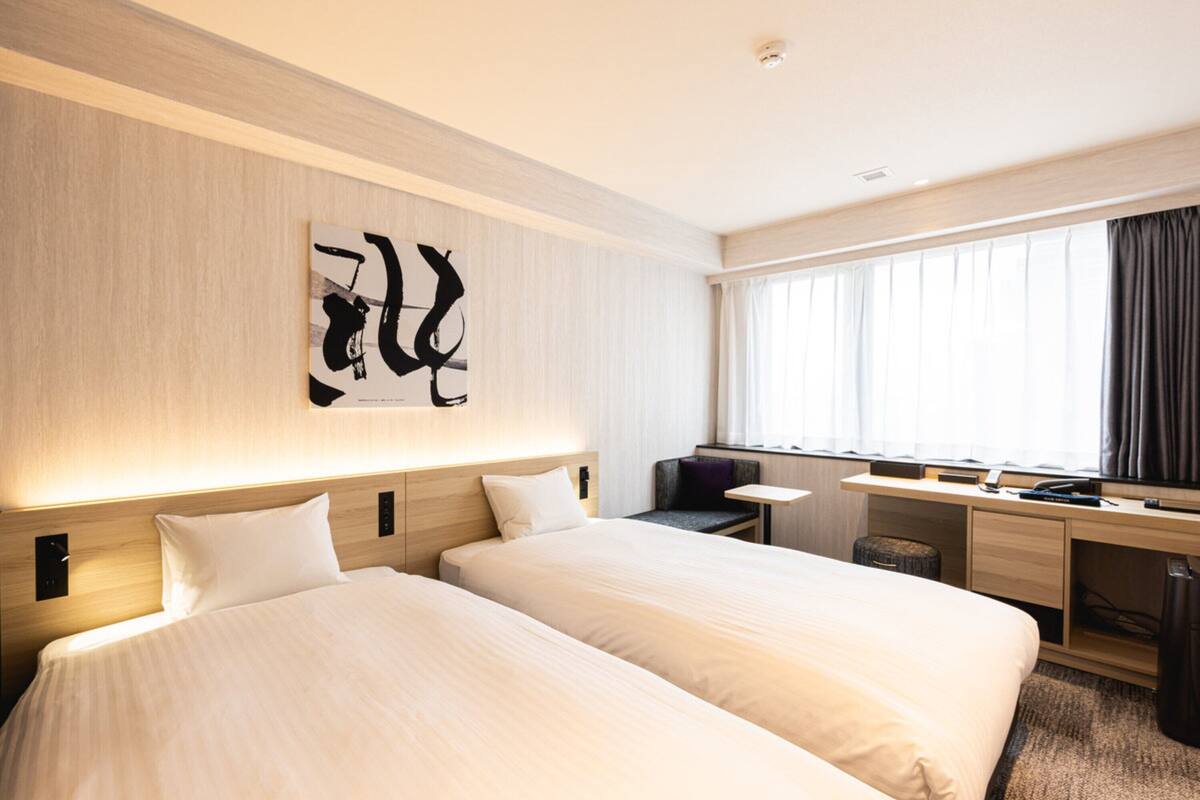 Habitación de hotel con dos camas, un escritorio, una silla y un ventanal con cortinas.