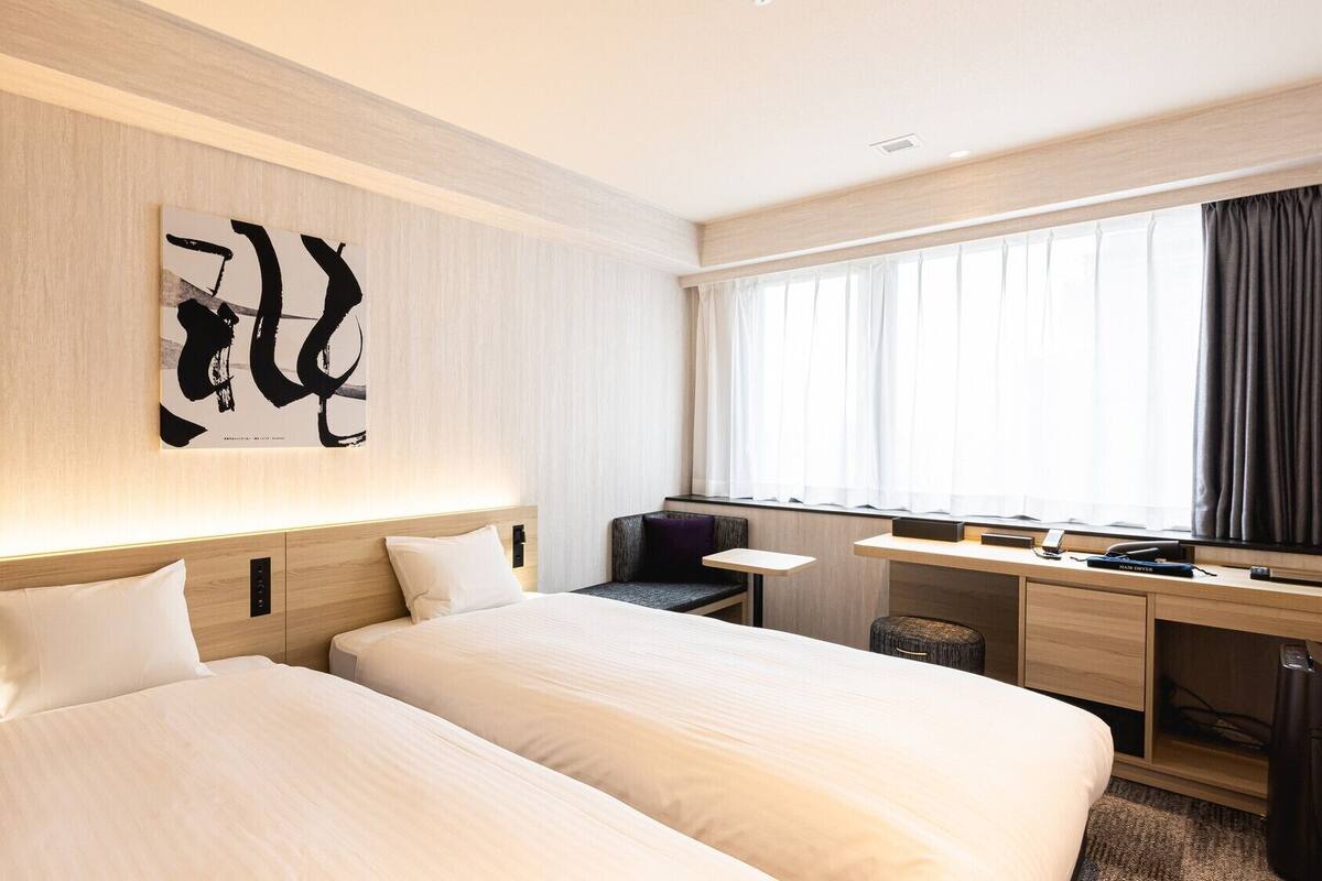 Habitación de hotel con dos camas, un escritorio, una silla, un ventanal con cortinas y una obra de arte en la pared.