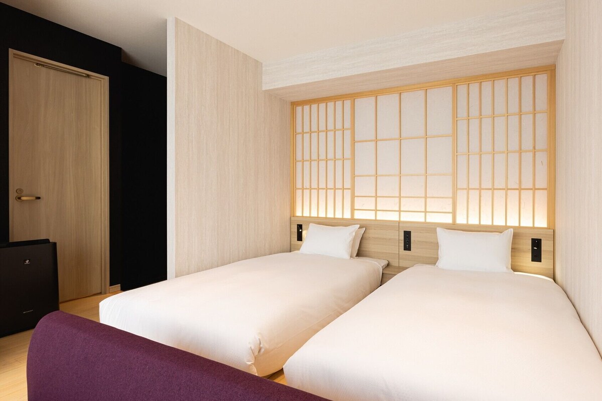 Habitación de hotel con dos camas, cabecero de madera con diseño de cuadrícula, armario de color oscuro y una banca púrpura.