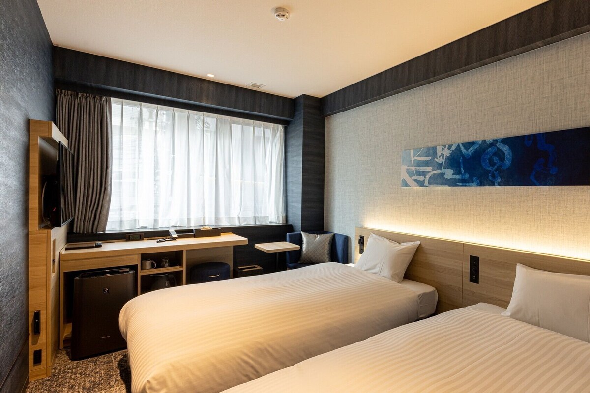 Habitación de hotel con dos camas, un escritorio, un televisor y un ventanal con cortinas.