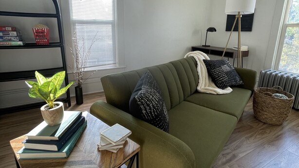Un salón con un sofá verde, una mesita con libros, una planta en maceta y una estantería con juegos de mesa y un bote de palomitas.
