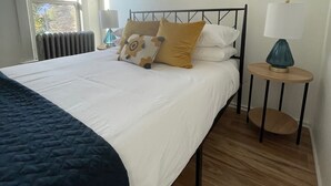 Una cama bien hecha con edredón blanco y dos almohadas, una mesita de noche con lámpara, y una ventana con vistas a árboles.